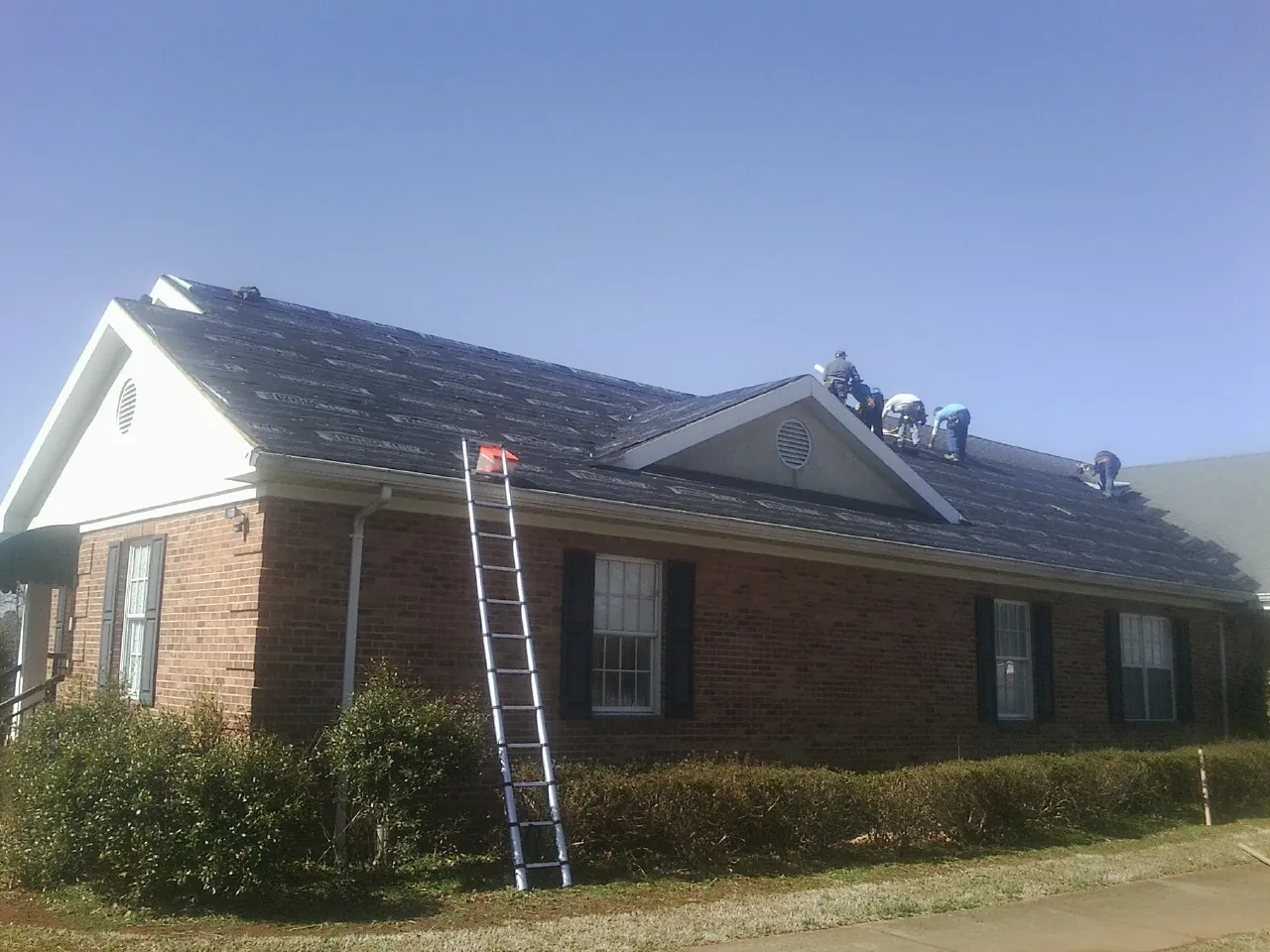 Crew tearing shingles off a brick ranch roof with a ladder set — Metal Roof Repair work by our crew in Rensselaer