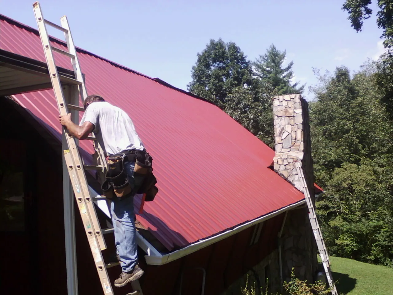 Roofer on a ladder beside a red metal roof and stone chimney — Soffit & Fascia Repair by Roofing Brothers in Rensselaer